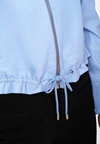 Helle blaue Lederjacke mit gerafft gesäumtem Saum, ausgestattet mit einem Metallreißverschluss und einer Kordelzugtaillierung mit silbernem Zubehör. Glatte Textur.