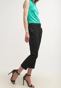 Top sans manches vert en tissu lisse, associé à un pantalon noir slim jusqu'aux chevilles. Des talons aiguilles noirs avec des brides autour de la cheville complètent la tenue.