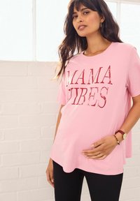 Roze t-shirt met korte mouwen en de tekst "MAMA VIBES" in het rood. Zachte katoenen stof, losse pasvorm, gedragen boven zwarte geribde leggings.