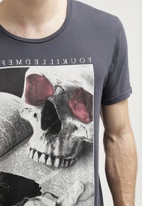 Tmavě šedé tričko s grafickým potiskem lebky s červenými očima na texturovaném pozadí s textem a stínovými prvky.