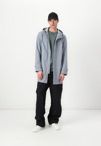 Lys grå hoodie-jakke over en grøn sweatshirt, parret med sorte cargo-bukser og hvide sneakers; afslappet, løs pasform med funktionelle detaljer.