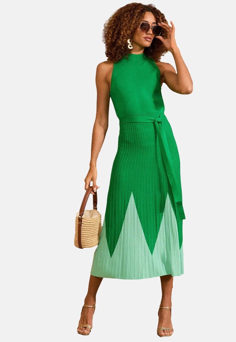 Abito verde a collo alto con design plissettato, che sfuma verso un orlo verde menta. Include una cintura coordinata e una borsa intrecciata di tessuto.