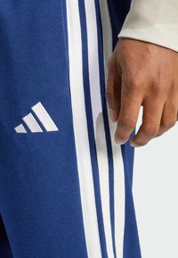 Pantalones de chándal azules con rayas blancas a lo largo de los lados. Presenta el logo de Adidas en blanco en la pierna izquierda y un primer plano de una mano agarrando la tela.