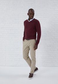 Pulover maron, cămașă albă cu guler, pantaloni krem și pantofi slip-on maro. Textură netedă, design ajustat, poziție în picioare lângă un perete din cărămidă albă.