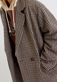 Cappotto houndstooth marrone e beige con revers a punta, due bottoni e tessuto testurizzato, indossato sopra un hoodie marrone con dettagli bianchi.