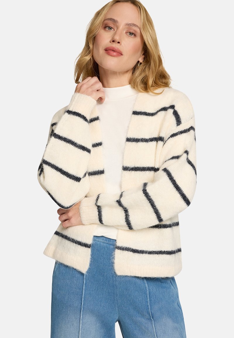 Cardigan crème avec rayures bleu marine foncé, texture douce, devant ouvert, manches longues et poignets côtelés. Associé à un haut blanc et un jean.