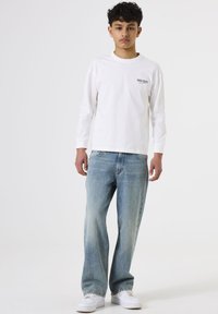 Witte katoenen t-shirt met lange mouwen en zwarte tekst op de borst, gecombineerd met lichtblauwe versleten spijkerbroek en witte sneakers.