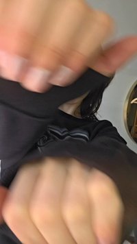 Osoba w czarnej koszulce z długim rękawem z uniesionymi pięściami blisko kamery, częściowo zasłaniająca twarz i górną część ciała.