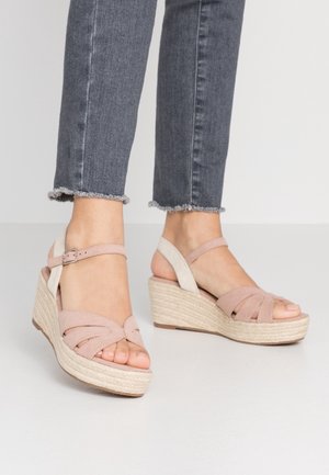 Füße in hellrosa Keilsandalen mit Knöchelriemen und geflochtenen Sohlen, getragen zu ausgefransten dunkelgrauen Jeans.
