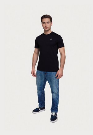 Ung mand iført en sort kortærmet t-shirt med logo, blå jeans og sorte sneakers, står foran en ensfarvet lys baggrund.