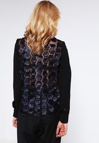 Svart blus med genomskinlig rygg och blå och svarta blommiga broderier, långa ärmar och knäppta manschetter.