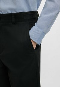 Pantaloni neri in tessuto liscio, con taglio dritto, tasche laterali e vita con logo. Indossati con una camicia azzurro chiaro.