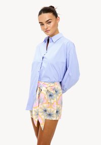 Mujer con un moño elegante que lleva una camisa de botones azul claro y pantalones cortos de envoltura con patrón floral en tonos pastel y un lazo atado, posando contra un fondo blanco.