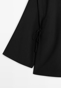 Détail d'un vêtement noir à manches longues avec un cordon en tissu noué à la couture latérale.