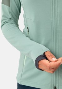 Hellgrüne Outdoor-Jacke aus strukturiertem Stoff, mit dunkelgrauen Stulpenakzenten, einer Reißverschlusstasche und einem vollen Frontreißverschluss.