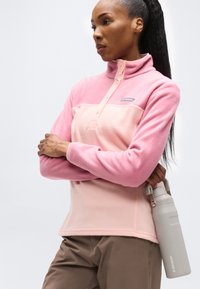 Γυναίκα με μακριά μαλλιά, φορώντας ροζ φλις πουλόβερ και καφέ παντελόνι, κρατώντας ένα γκρι μπουκάλι νερού στο πλευρό της, με λευκό φόντο.