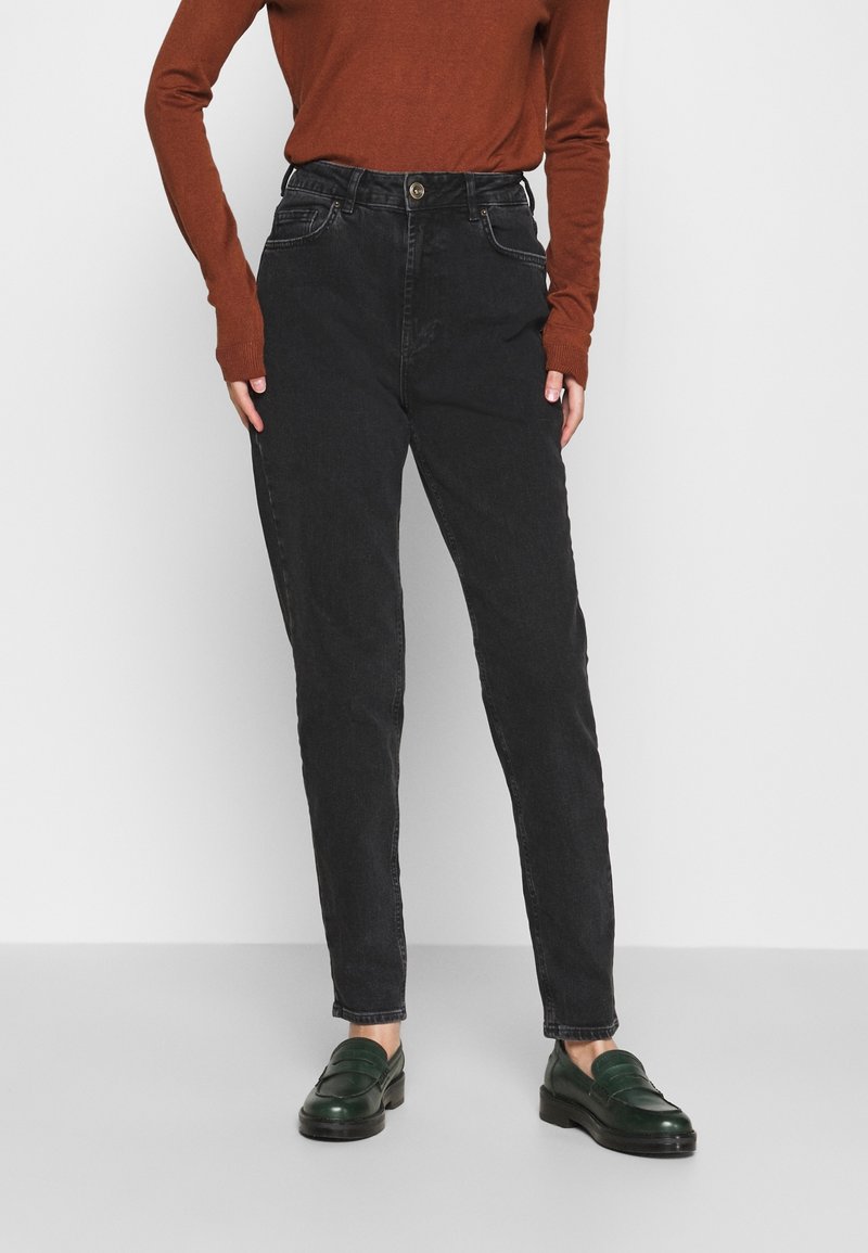 Personne portant un jean noir taille haute, un haut marron à manches longues et des mocassins en cuir vert foncé, debout sur un fond gris clair.