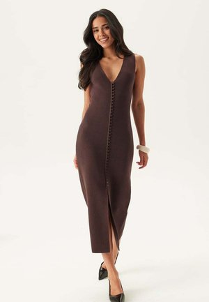 Vrouw glimlachend, draagt een mouwloze donkerbruine midi-jurk met knopen aan de voorkant en een voorsplit, met zwarte puntige hakken en een witte armband om haar pols.