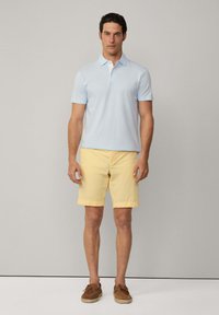 Světle modré polo tričko, krátké rukávy a límeček; doplněné o žluté šortky. Hnědé mokasíny se šněrováním celý outfit dotvářejí. Celkově hladká textura.