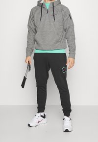 Haut à capuche zippé gris avec cordons de serrage noirs, associé à un pantalon de jogging noir avec une accent graphique. Des sneakers blanches complètent la tenue.