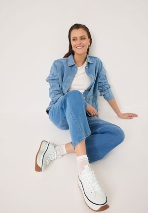 Veste en denim et pantalon assorti en bleu clair, associés à des baskets blanches avec une semelle en caoutchouc. Modèle assis contre un fond neutre.