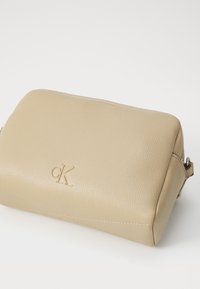 Beige lærpung med en teksturert overflate, preget med logoen "ck" på forsiden og med glidelås på siden.