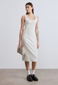 Φωτεινό γκρι, ριμπ φόρεμα με τιράντες και στρογγυλή λαιμόκοψη. Το μοντέλο κρατά μια μικρή μπεζ τσάντα χειρός. Φορούν μαύρα παπούτσια με πάτο και λευκές κάλτσες.