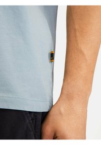 Lyseblå bomulds T-shirt med sideskæring og et sort og gult logo mærke nær kanten, der viser en tæt på arm og stoffets tekstur.