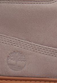 Gros plan sur du cuir suédé beige avec une couture cousue et un logo d'arbre embossé au-dessus du bord en caoutchouc beige.