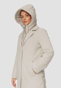 Beige wollen blendjas met een puffer-achtige, gewatteerde capuchon, ritssluiting aan de voorkant en zijzakken. Getailleerde pasvorm met een strakke afwerking en een gestructureerde stof.