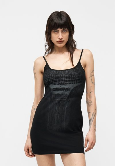 Femme aux cheveux foncés portant une mini robe noire sans manches avec un motif abstrait et des bretelles fines, montrant des tatouages sur les bras, sur un fond uni.