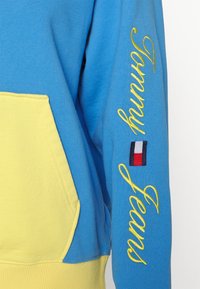 Manche de sweat à capuche bleue et jaune avec une broderie jaune "Tommy Jeans" et un petit drapeau logo Tommy Hilfiger rouge, blanc et bleu.