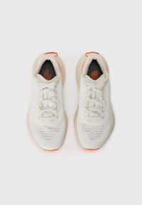 Pár bílých a světle šedých atletických tenisek s oranžovými detaily, s tkaničkami, umístěnými vedle sebe na bílém pozadí.