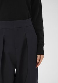 Pantaloni a plissettatura blu navy con una vita alta, realizzati in un tessuto liscio. La parte superiore è un maglione nero in maglia con polsini a costine.