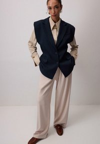 Námornícky oversized vestový kabát s ramennými vypchávkami, béžová košeľa na gombíky s perlami, široké krémové nohavice, doplnené hnedými mokasínami.