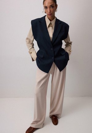 Marineblå oversized vest med skulderpuder, beige skjorte med perleaccentuer, og brede cremefarvede bukser, suppleret af brune loafers.