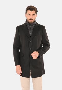 Uomo con barba che indossa un cappotto nero con bottoni sopra una giacca nera con cerniera e pantaloni beige, in piedi davanti a uno sfondo semplice.
