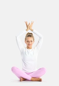 Fehér, hosszú ujjú felső és rózsaszín leggings, keresztbe tett lábakkal ül, kezét a feje fölé emeli, szeme csukva, bőre sima textúrájú.