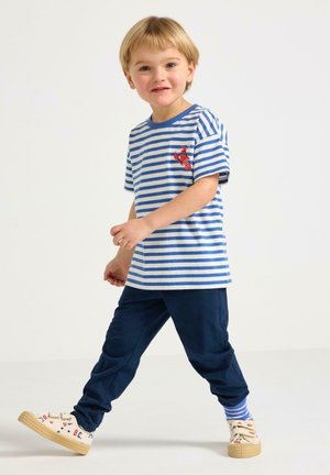 Nuori poika kävelee, hänellä on päällään sinisävyinen raidallinen t-paita, jossa on hummereiden kuvio, tummansiniset housut, raidalliset sukat ja beiget lenkkarit, joissa on punaisia ja sinisiä kirjaimia.