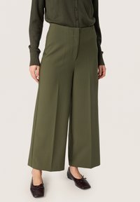 Olivgrüne weite Hose aus glattem Stoff, mit einem Vorderbügel und hoher Taille. Kombiniert mit dunklen, strukturierten Ballerinas.