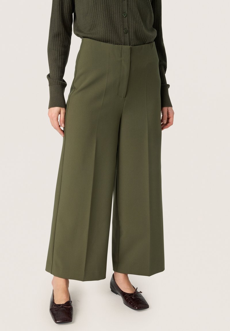 Olivgrüne weite Hose aus glattem Stoff, mit einem Vorderbügel und hoher Taille. Kombiniert mit dunklen, strukturierten Ballerinas.