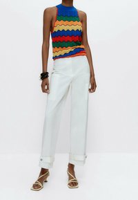 Vrouw in een mouwloze top met een kleuren zigzagpatroon, witte wijde broek met omslagen, beige strappy hakken en zwarte armbanden om één pols.
