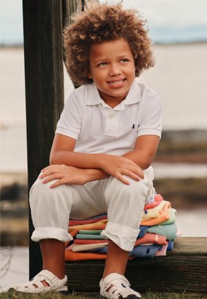 Παιδί με σγουρά μαλλιά που φοράει λευκή πόλο και παντελόνι, κάθεται σε ξύλινο παγκάκι στοιβαγμένο με διπλωμένα πολύχρωμα ρούχα, έξω κοντά στο νερό.