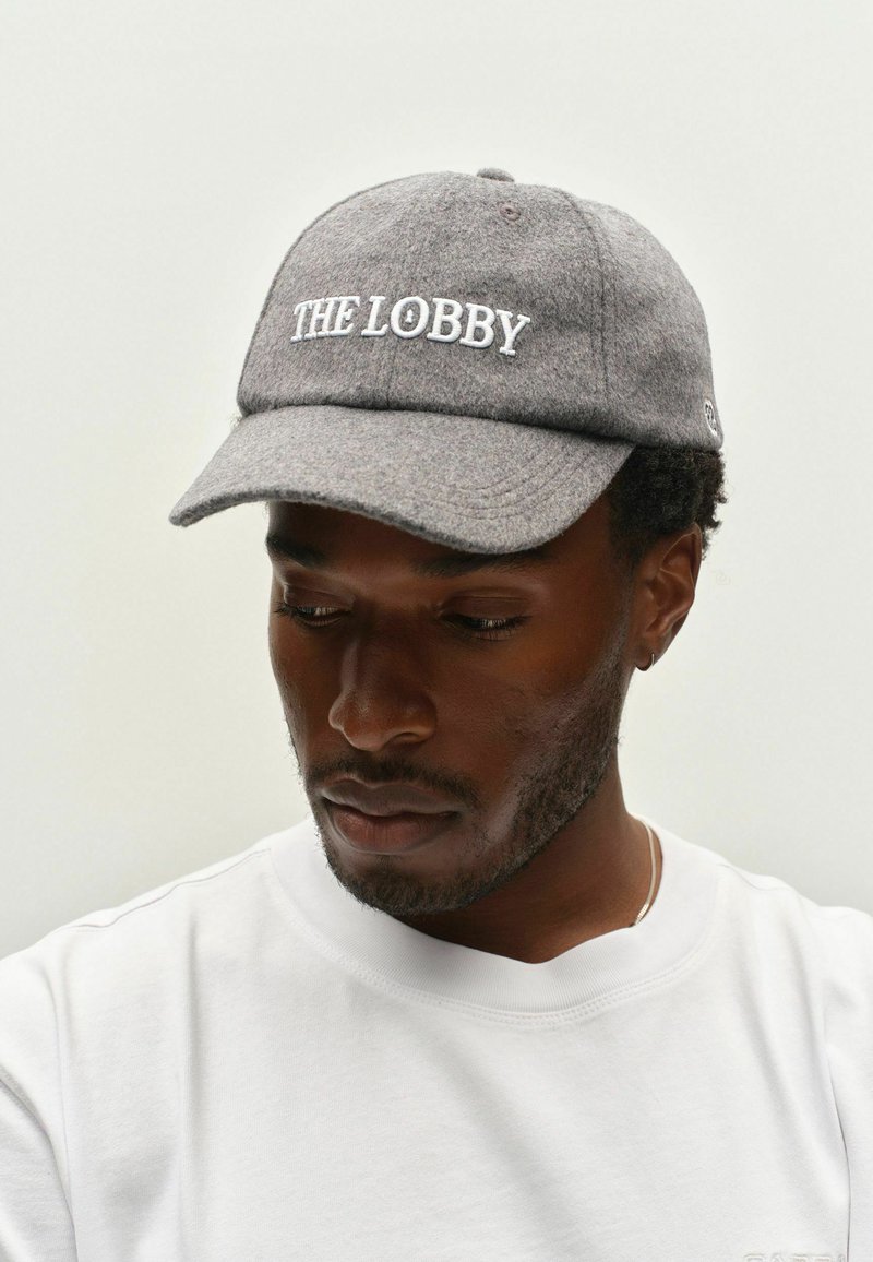 Homme à la peau foncée portant une casquette grise avec le texte "THE LOBBY" et une chemise blanche, regardant vers le bas sur un fond uni.