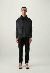 Giacca bomber navy con tasche laterali, colletto alto e zip frontale. Abbinata a pantaloni neri e sneakers beige. Texture liscia.