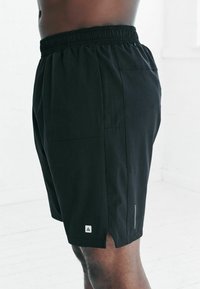 Juodi sportiniai šortai su elastinga juosmens juosta, šoninėmis kišenėmis ir tinklelio detalėmis šonuose. Prie apačios yra nedidelis baltas logotipas.