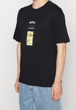 Mężczyzna w czarnej koszulce z krótkim rękawem z beżowym napisem „BOSS AJBXNG” oraz abstrakcyjnym wzorem, w połączeniu z jasnoniebieskimi jeansami.