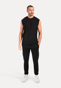 Schwarzes ärmelloses Top und schwarze tapered Sweatpants, beide aus glattem Stoff, kombiniert mit weißen Schnürsneakern. Minimalistisches Design.