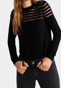 Haut noir à manches longues avec un motif rayé transparent sur les épaules. Le tissu semble doux, avec un col rond et des poignets ajustés, porté avec un jean foncé.