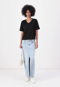 Maglietta nera a V abbinata a una gonna di jeans azzurro chiaro con spacco frontale. La modella tiene in mano una piccola borsa bianca; indossa sneakers bianche.
